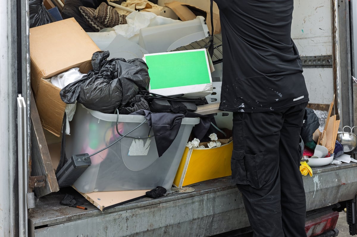 Worker unloading mixed waste, household items, and rubbish from the rear of a commercial truck, showing waste removal, clearance work, recycling, and manual handling tasks